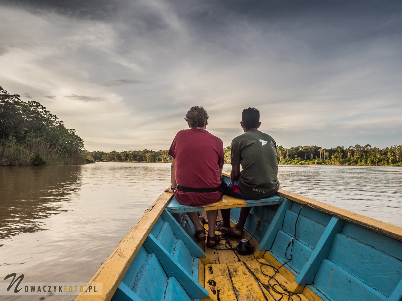Dwóch panów siedzi na czubku łodzi i patrzy przed siebie na dżunglę i rzekę